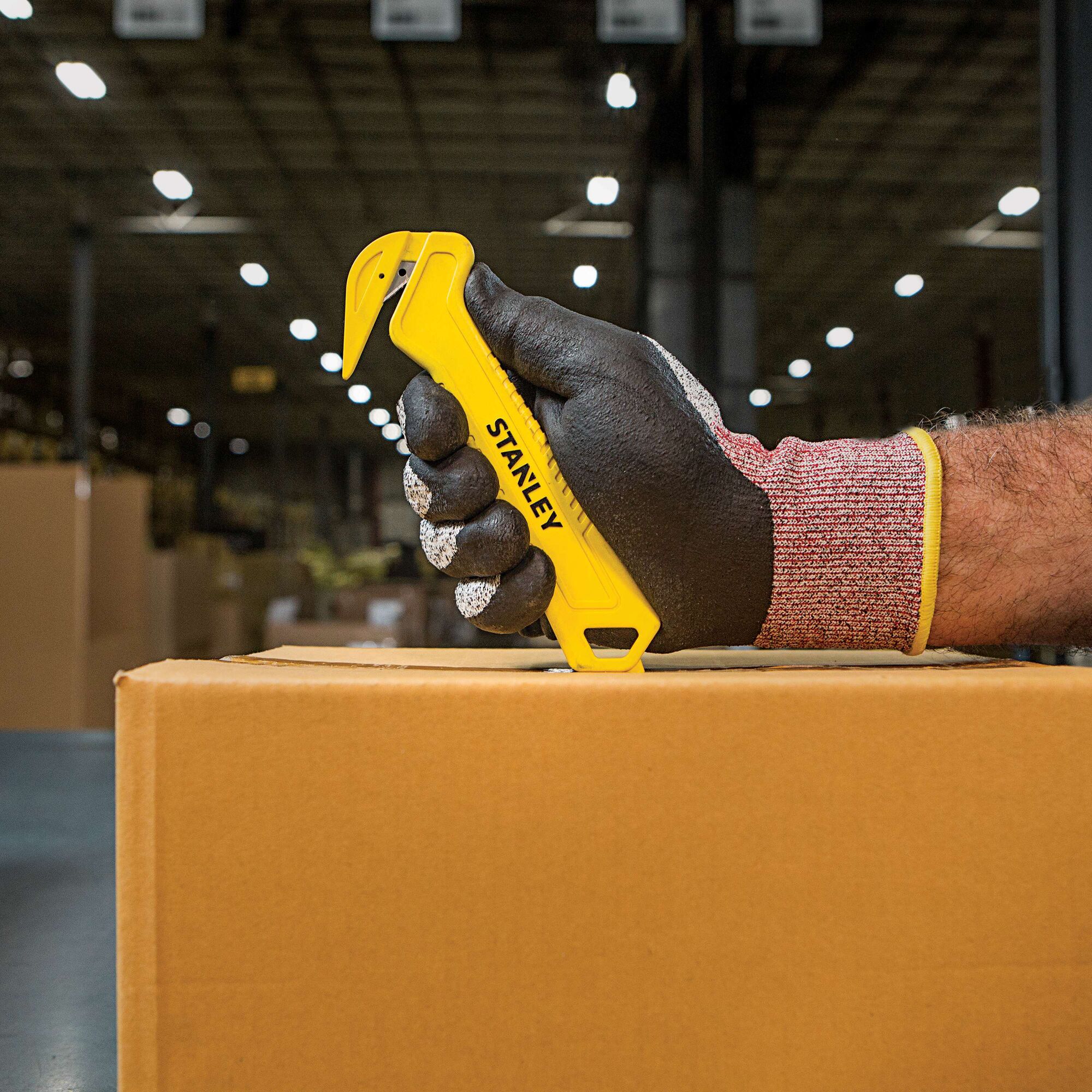 Stanley STHT10355 safety knife in gloved hand cutting cardboard box in warehouse setting.