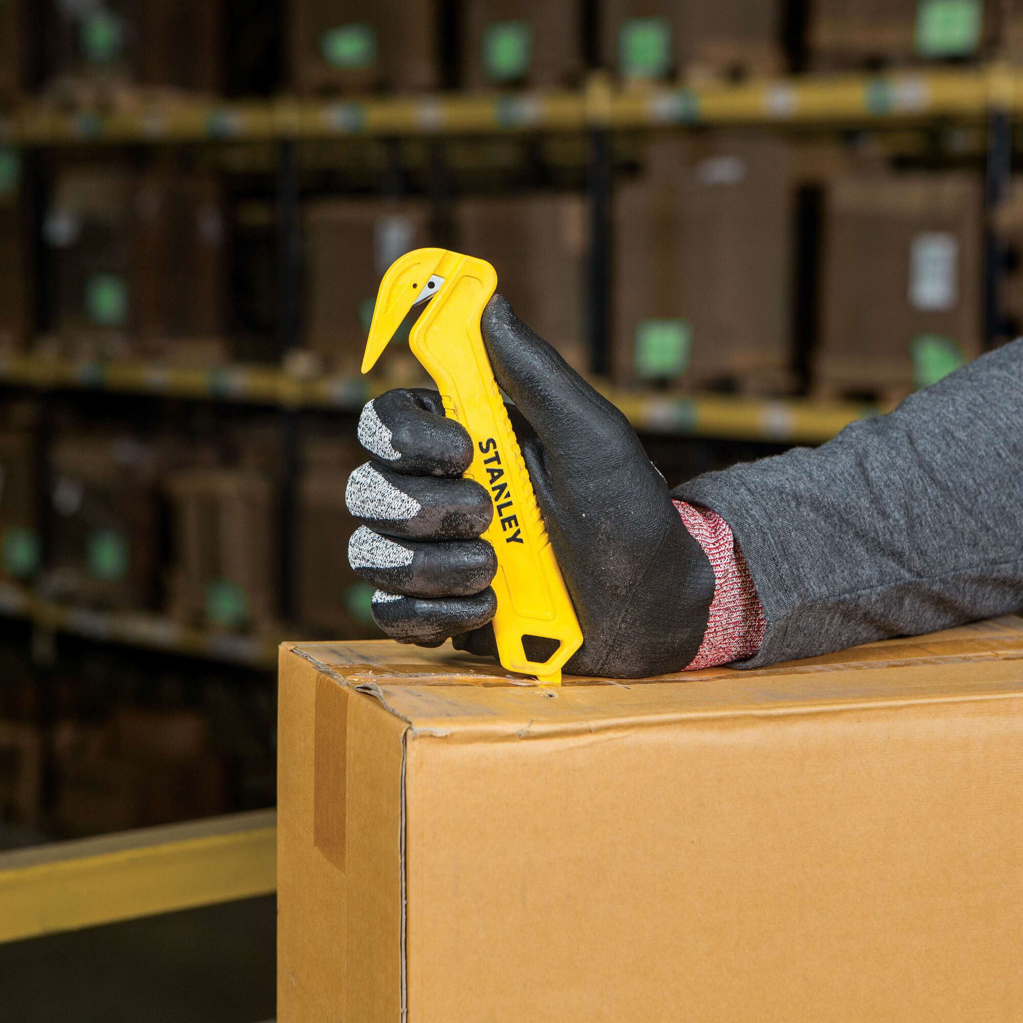 Stanley STHT10355 yellow safety box cutter held by gloved hand opening cardboard box in warehouse.