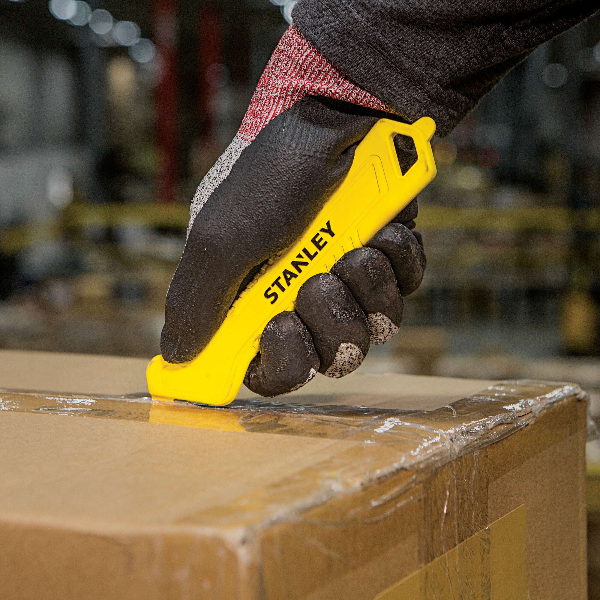 Stanley STHT10355 safety utility knife cutting tape on a cardboard box, close-up in warehouse setting.
