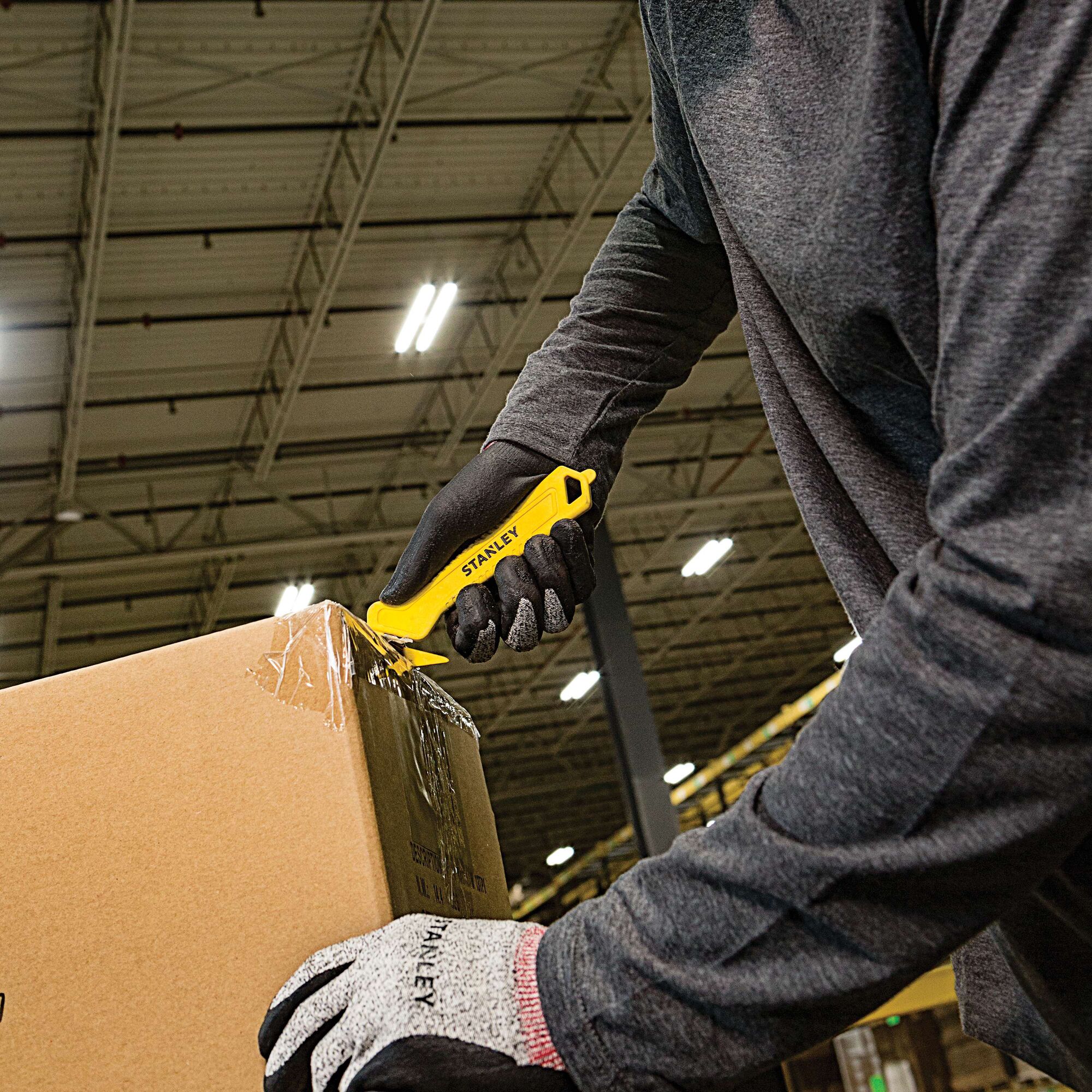 Photo of person using STANLEY STHT10355 yellow safety knife to open a cardboard box in a warehouse setting.