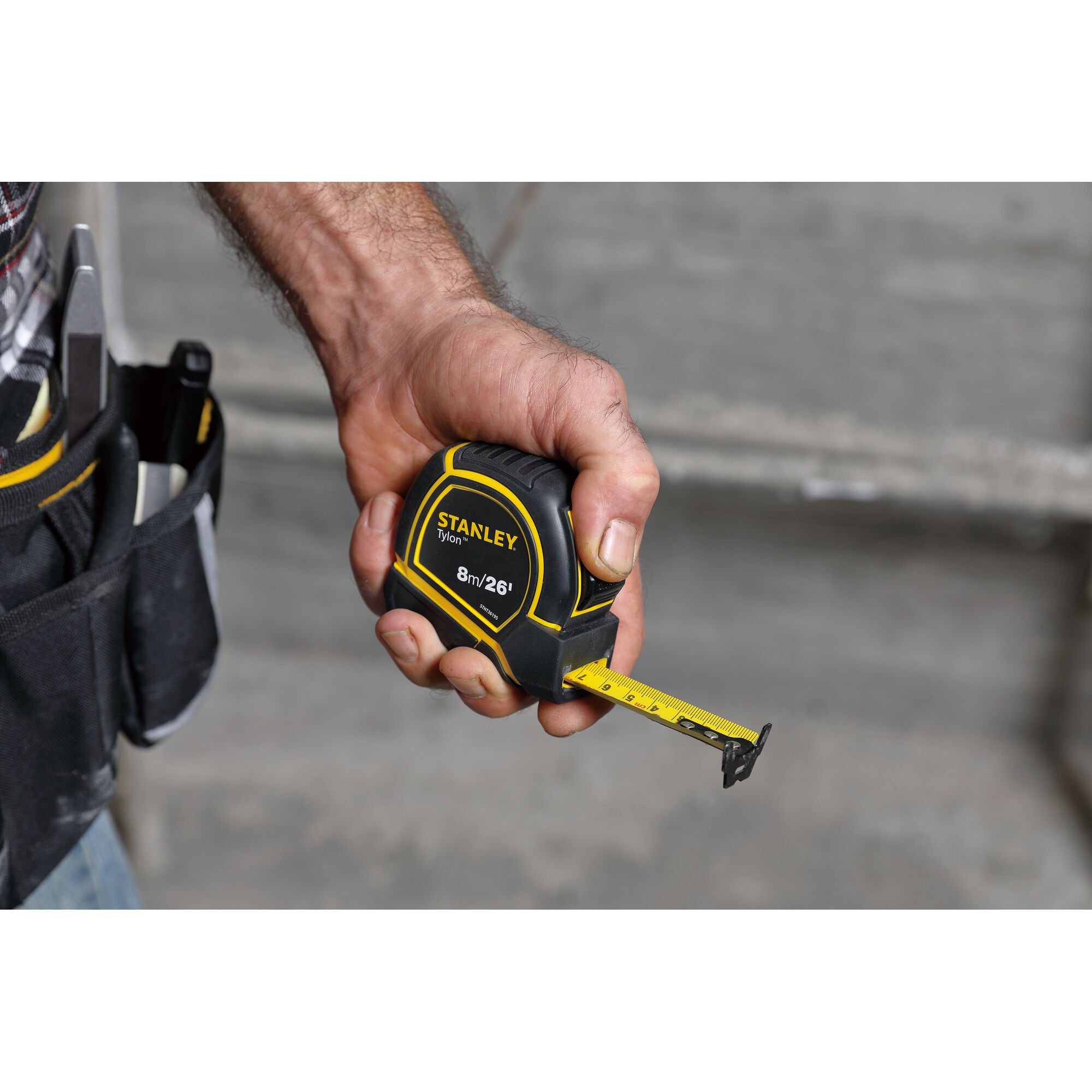 Stanley STHT36195 tape measure in black and yellow, held in hand, with belt pouch and tools visible.
